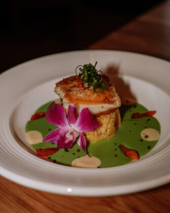 Seared fish served over a composed base with bright green sauce, garnishes, and an edible orchid.
