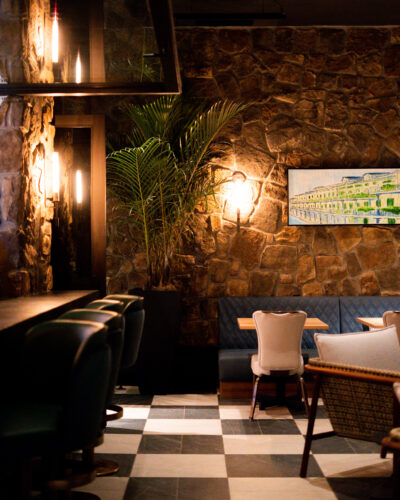 Moody restaurant interior with stone walls, checkerboard flooring, bar seating, and warm lighting.