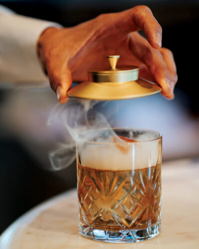 Smoked amber cocktail revealed beneath a gold-topped cloche in a cut-crystal glass.