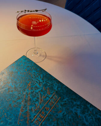 Red cocktail garnished with a sprig of thyme beside an Aqua Beef & Reef menu on a white tablecloth.