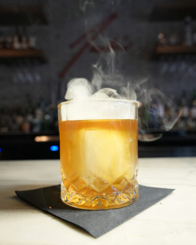The Soft N' Smokey cocktail sits on the counter as smoke drifts off. 