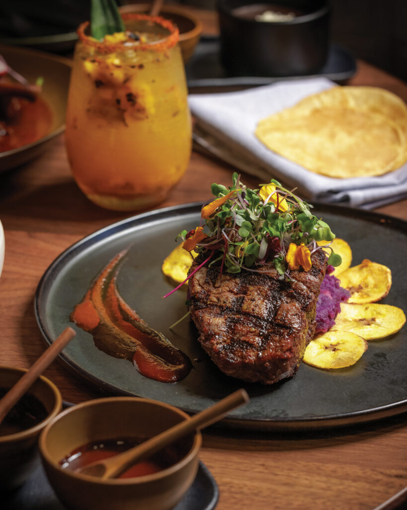 Grilled steak topped with microgreens and edible flowers, served with plantains and colorful sauces.