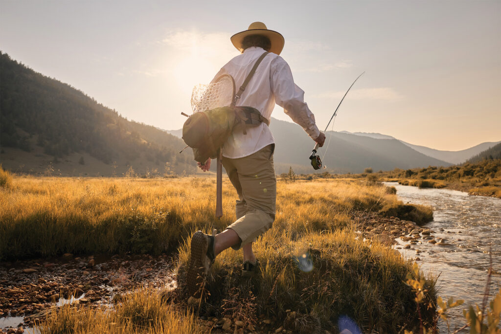 A fly fisherman carrying a rod and fishing gear walks along a mountain stream at sunset.