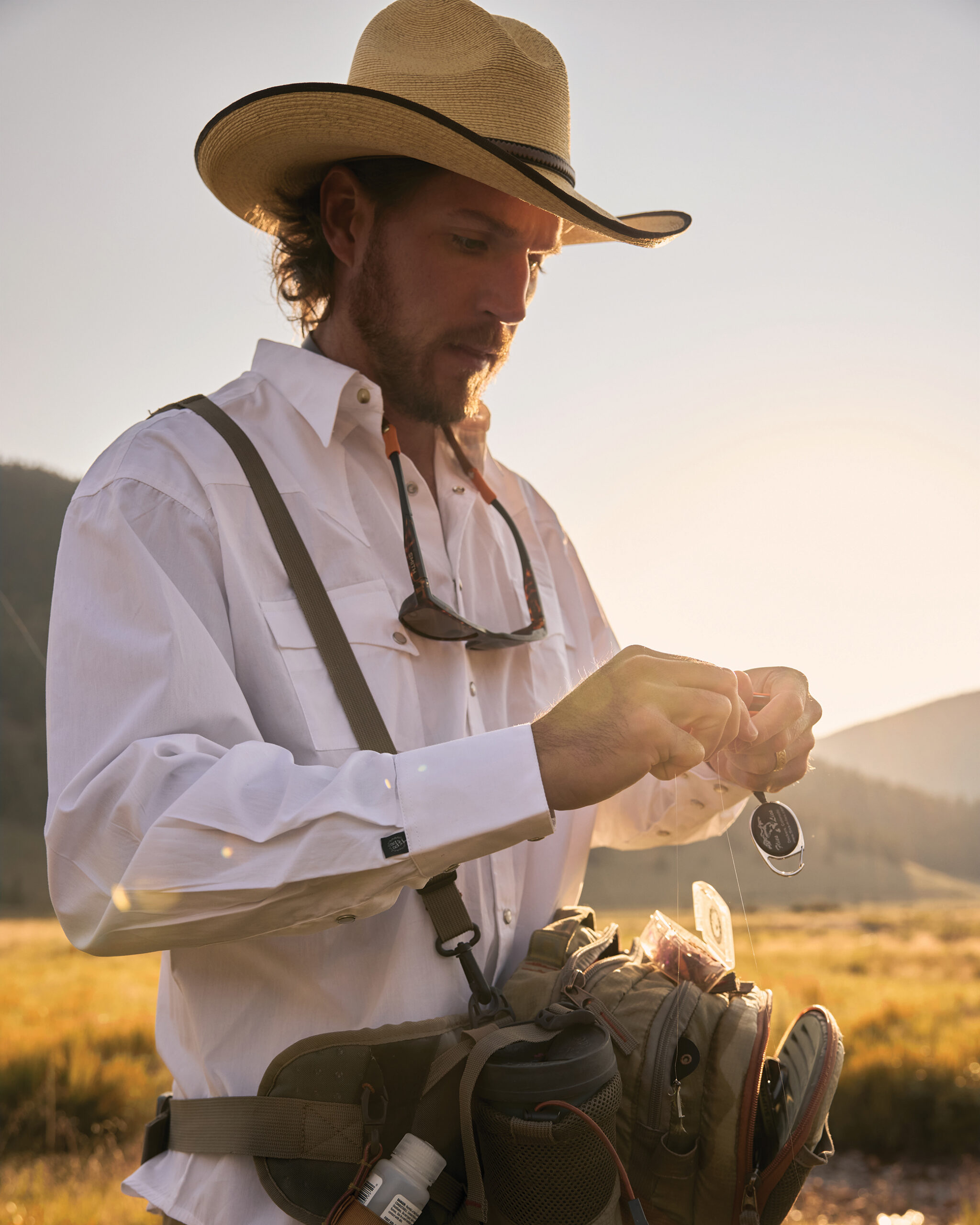 A man in a cowboy hat prepares his fly-fishing line beside a mountain stream in golden evening light.