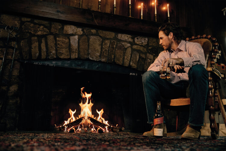 A man sits beside a stone fireplace with a drink in hand inside a rustic, candlelit lodge.