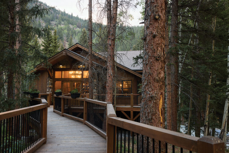 A wooden lodge nestled among tall pine trees is reached by an elevated wooden walkway.