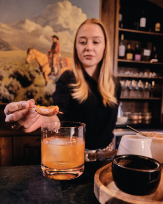 A bartender garnishes a cocktail with a citrus peel inside a rustic Western-style bar.