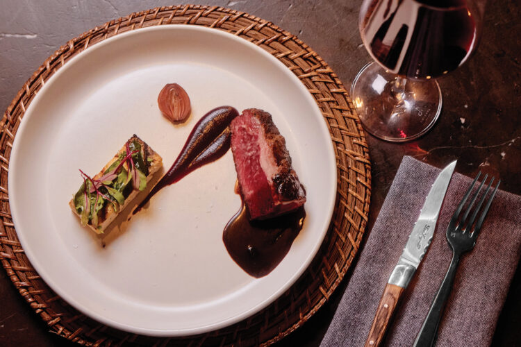 A plated lamb dish with sauce and a vegetable and polenta accompaniment sits beside a glass of red wine.