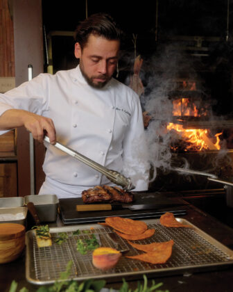 Chef Alex uses tongs to prepare a grilled steak in front of a wood-fired grill.