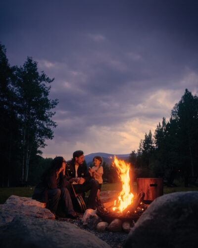A family gathers around a glowing outdoor fire pit at dusk with mountains visible beneath a dramatic sky.
