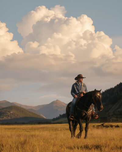 A cowboy on a black horse pauses in a sunlit mountain meadow beneath towering clouds.