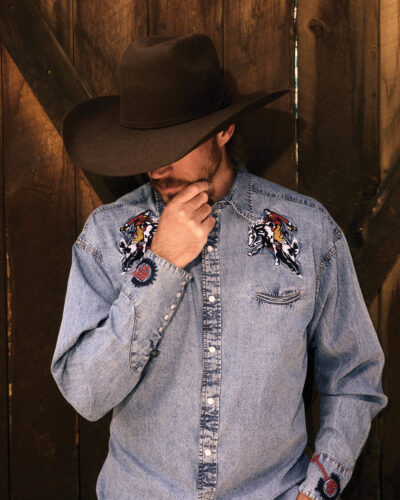 A man in a black cowboy hat and embroidered denim Western shirt from Rockmount Ranch Wear stands against a rustic wooden wall.