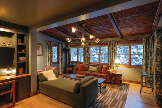 Renovated sitting room at Tumbling River Ranch featuring exposed wood beams, large windows overlooking snowy trees, patterned curtains and comfortable lounge seating.