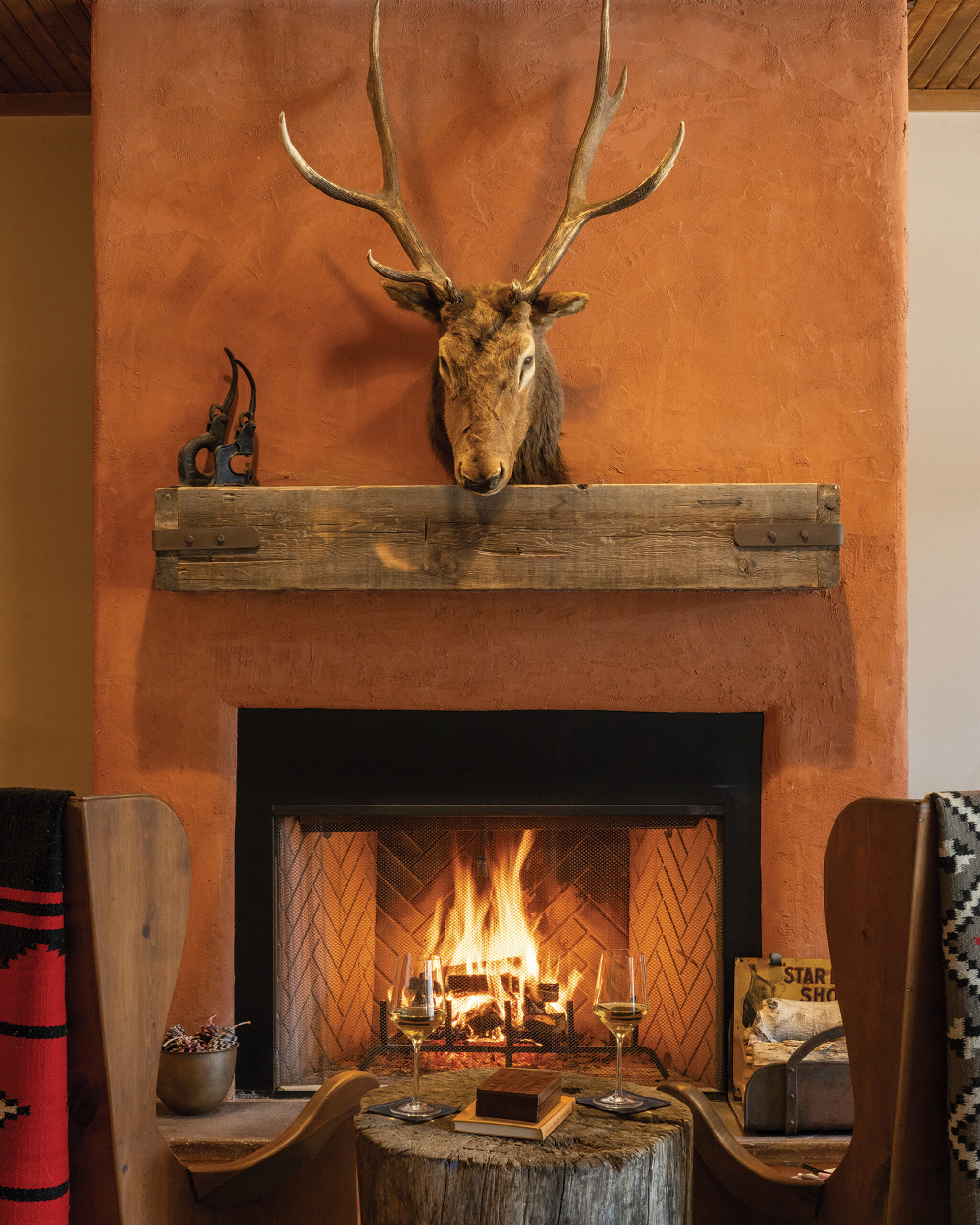 Fireplace at Tumbling River Ranch featuring a mounted elk above a roaring fire, rustic wood chairs, Southwestern blankets and wine glasses.