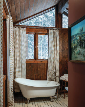 Clawfoot bathtub in a rustic Tumbling River Ranch bathroom surrounded by wood paneling, patterned curtains, Western artwork and snowy forest views.