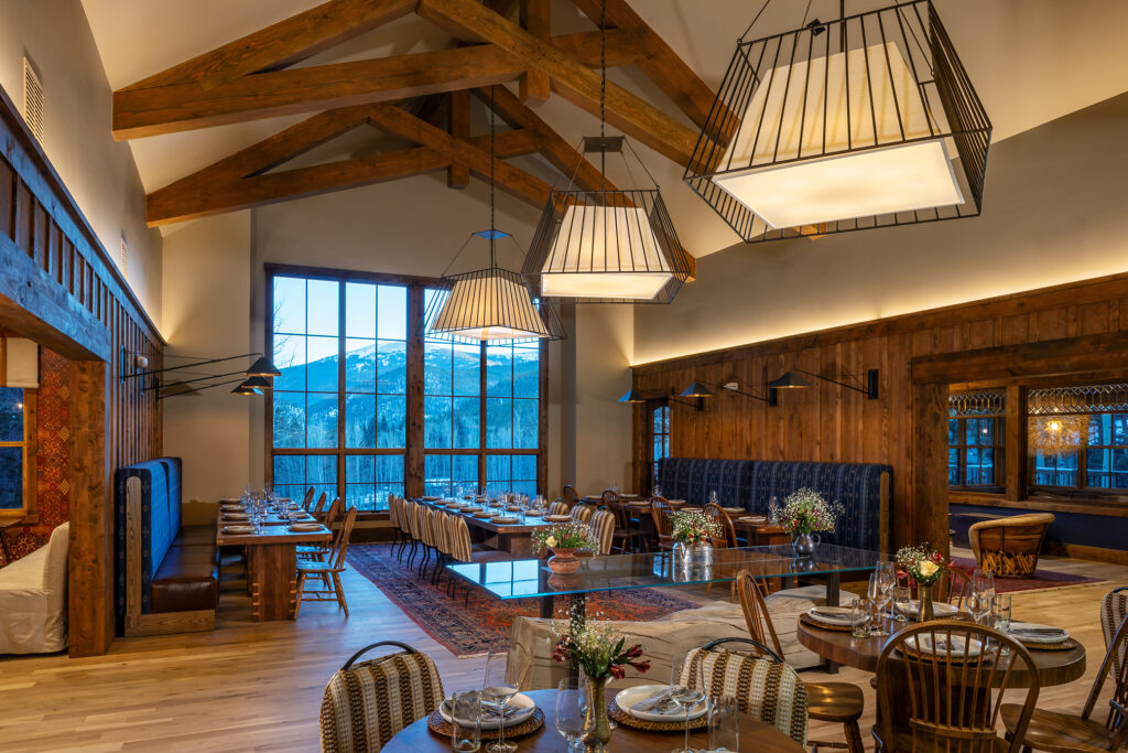 Dining room at Tumbling River Ranch with exposed timber trusses, oversized pendant lights, wood tables, patterned seating and mountain views through floor-to-ceiling windows.