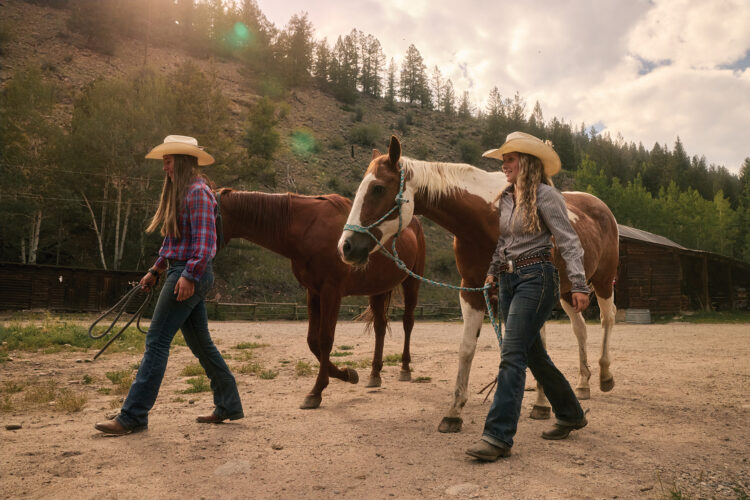 Two cowgirls in Western attire lead horses through a rustic mountain ranch.