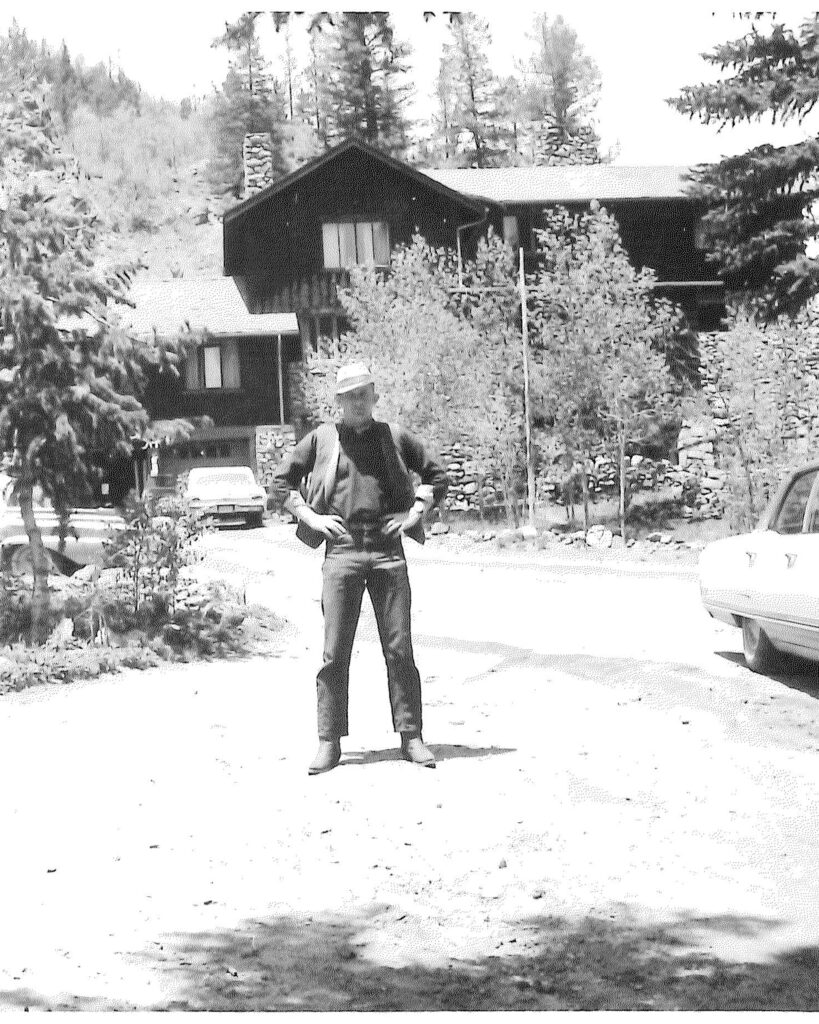 Historic black-and-white photograph of a cowboy standing in front of the stone-and-wood Ranch House at Tumbling River Ranch in Colorado.
