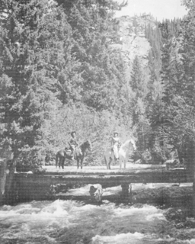 Historic black-and-white photograph of horseback riders crossing a bridge over a rushing creek near Tumbling River Ranch in the Colorado mountains.