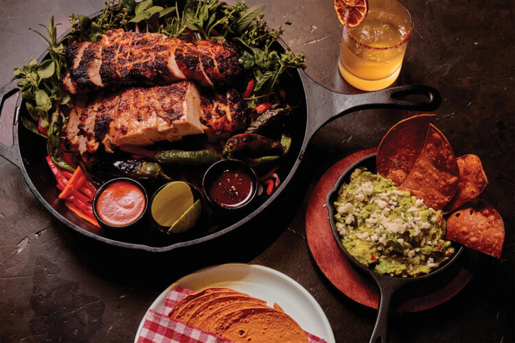 A spread of grilled meat, roasted peppers, guacamole, tortillas, sauces, and a cocktail arranged on a dark table.