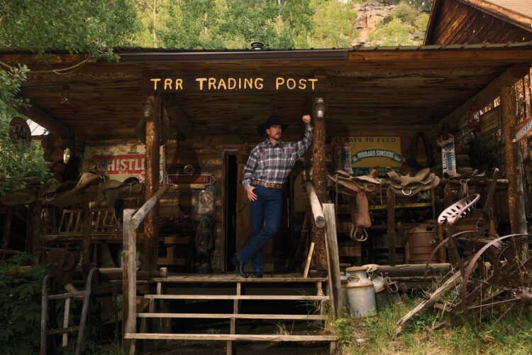 A cowboy stands on the porch of the Tumbling River Ranch Trading Post, surrounded by saddles, antique signs, and rustic ranch equipment.