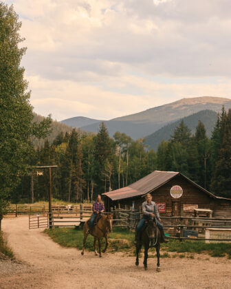 Two horseback riders travel past a rustic ranch barn with forested Colorado mountains rising in the background.