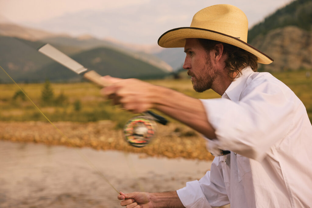 A man in a cowboy hat casts a fly-fishing line beside a mountain stream.