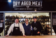 Three men posing behind a butcher counter in front of Tony’s Meats & Market’s dry-aged meat display.