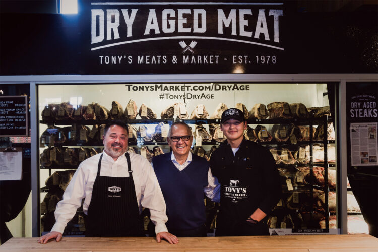 Three men posing behind a butcher counter in front of Tony’s Meats & Market’s dry-aged meat display.