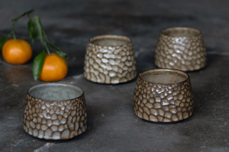 Four ceramic cups from Twisted Pine Pottery with pebble-like textures beside oranges.
