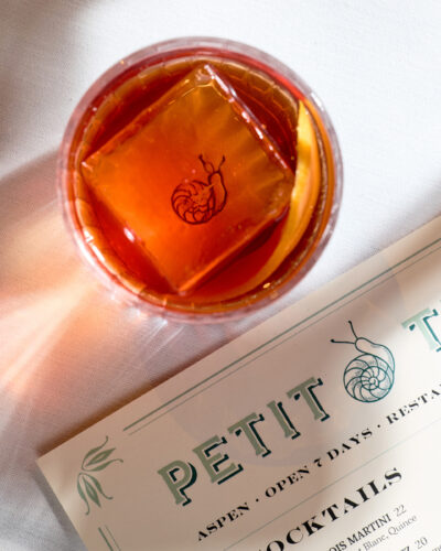 Amber cocktail with an orange peel and a snail-stamped ice cube beside a Petit Trois Aspen menu.