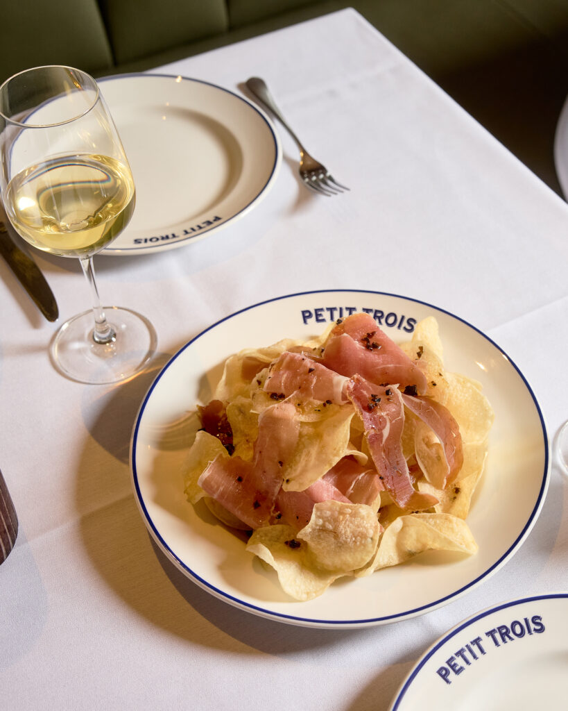 Potato chips topped with thinly sliced cured ham beside a glass of white wine at Petit Trois Aspen.