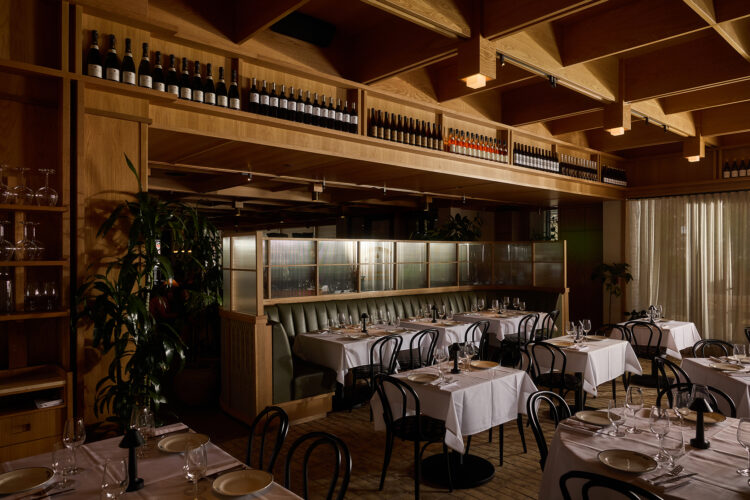 Petit Trois Aspen’s dining room with white tablecloths, green banquette seating, and shelves of wine.