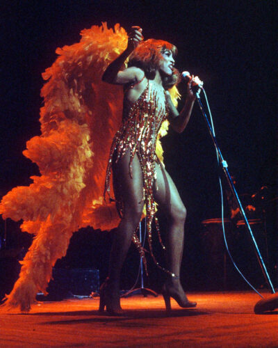 Singer performs in a glittering fringed costume with a dramatic orange feather cape.