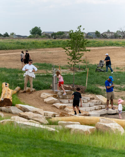 Families explore an outdoor play area with rocks and a water pump at Anythink Nature Library.