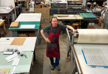 Open Press and Denver’s Printmaking Legacy Printmaker in a red apron stands beside a printing press in a studio filled with tools and worktables.
