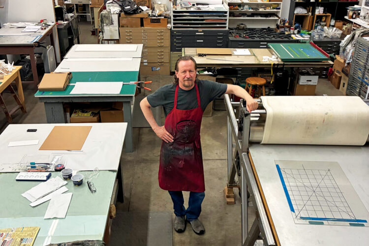 Printmaker in a red apron stands beside a printing press in a studio filled with tools and worktables.