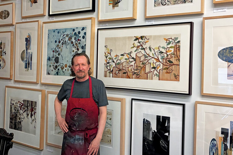 Artist in a red apron stands before a wall of framed abstract prints.
