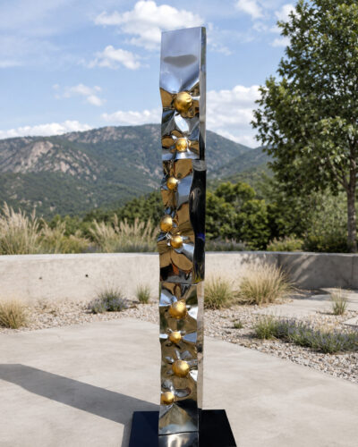 Reflective metal sculpture with gold spheres stands outdoors against a mountain backdrop.