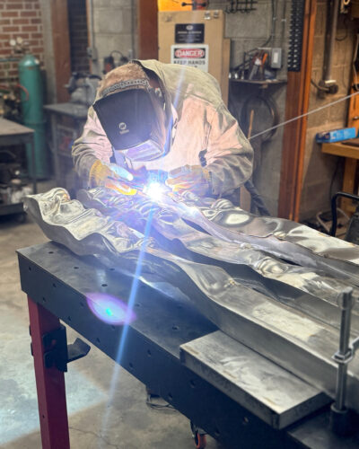 Sculptor in protective gear welds flowing metal forms on a workshop table.