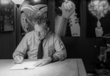 John Atencio Marks 50 Years of Jewelry Design Black and white photo of John Atencio drawing at a desk surrounded by artwork.