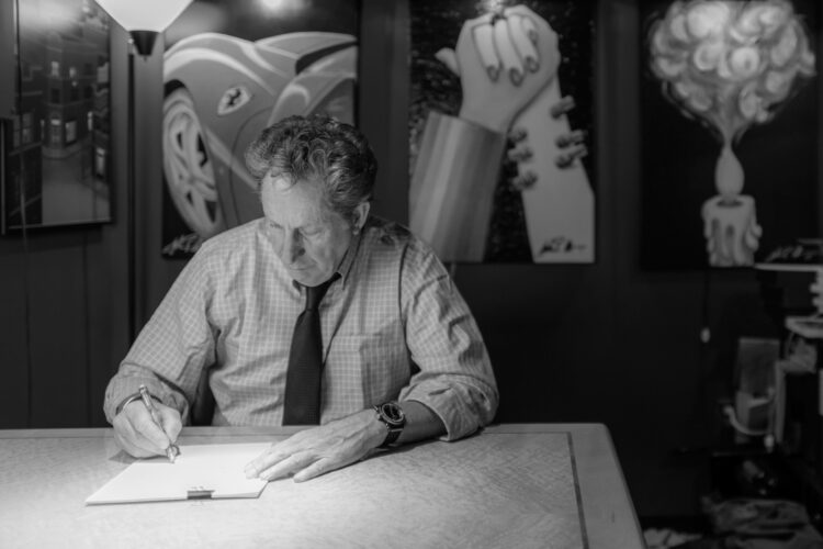 Black and white photo of John Atencio drawing at a desk surrounded by artwork.