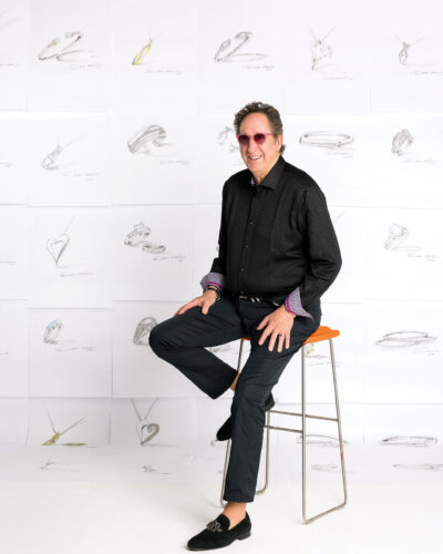 John Atencio seated on a stool in front of a wall of his jewelry sketches.