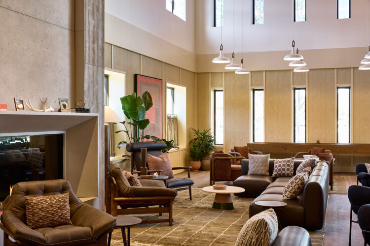 East & Co.’s lounge features leather seating, patterned pillows, a fireplace, and tall windows beneath pendant lights.