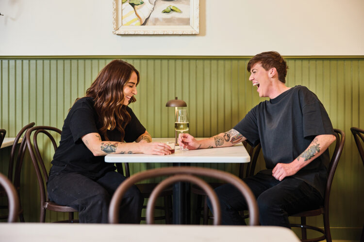 Two people laugh over drinks at a table against Mother Other’s green paneled wall.